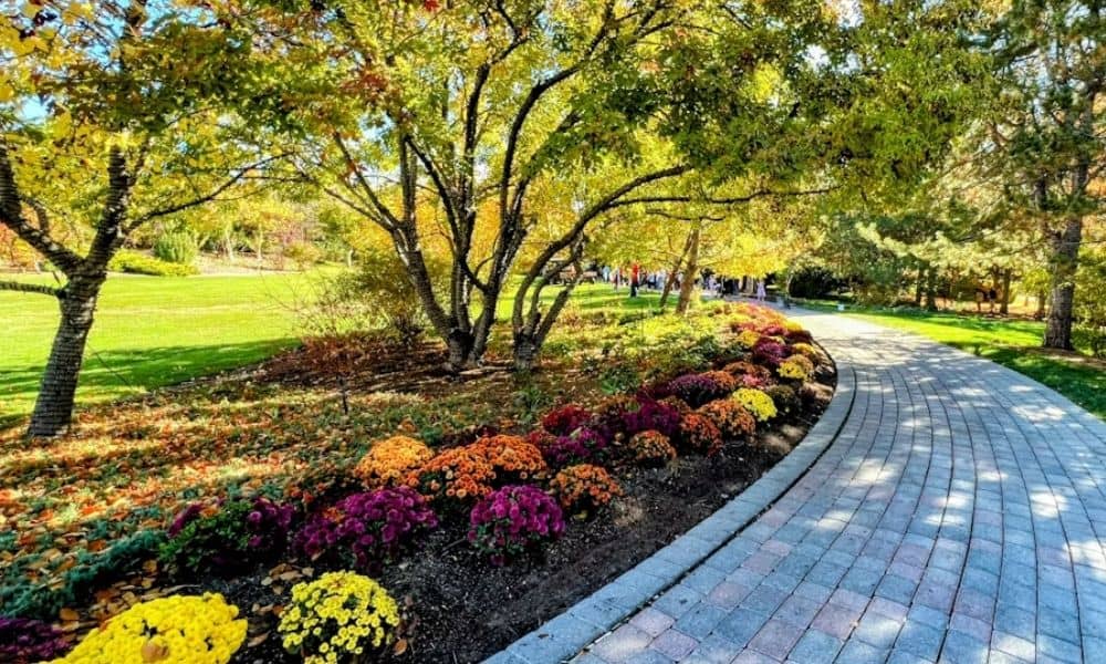 Curved masonry walkway winding through a landscaped yard with colorful flower beds, trees, and lawn