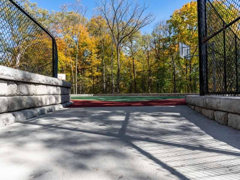 Stone retaining wall beside a backyard sport court with paver walkway, drainage area, and landscaped yard.
